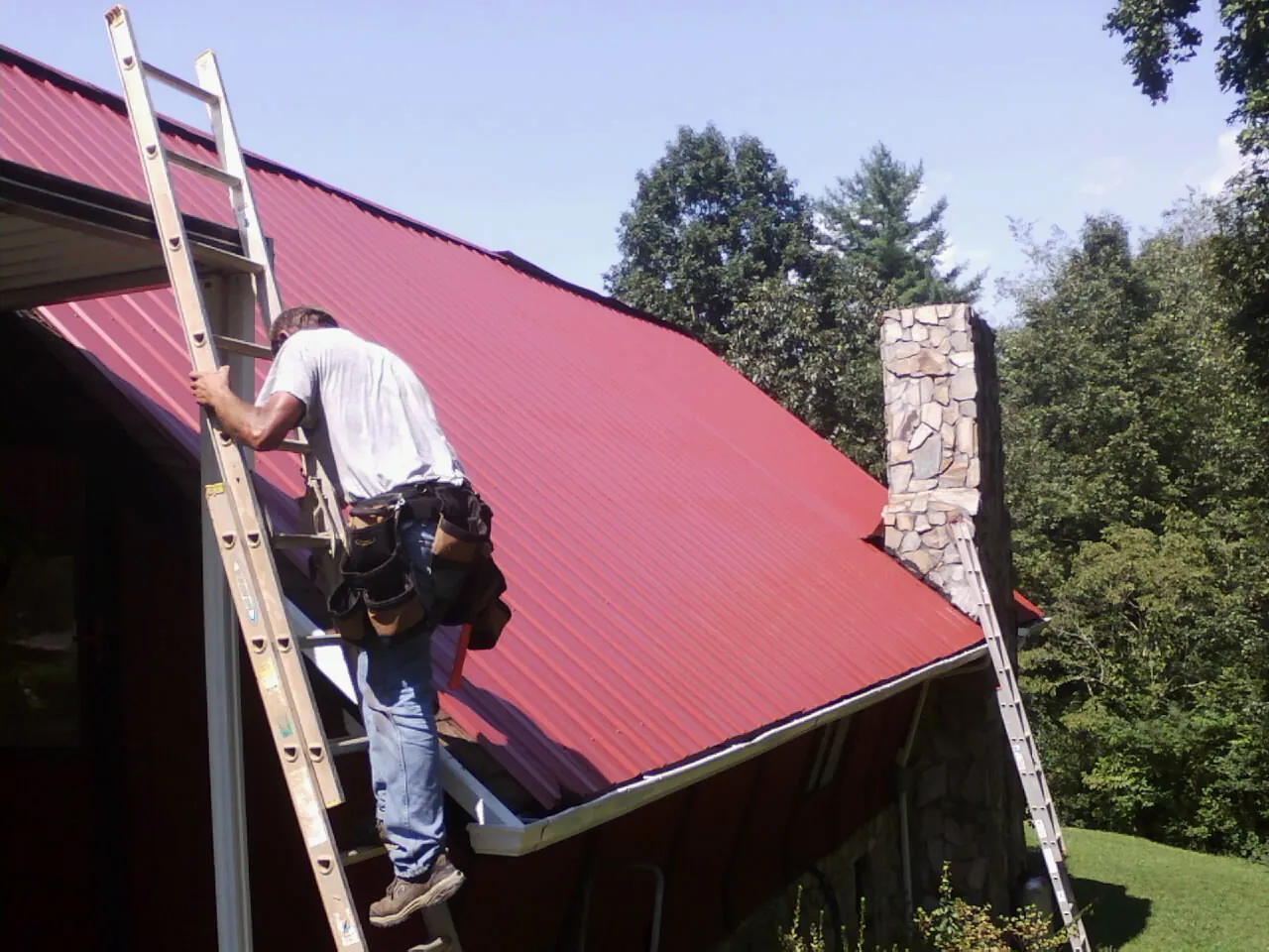 Roofer on a ladder beside a red metal roof and stone chimney — Soffit & Fascia Repair by Roofing Brothers in Lookout Mountain